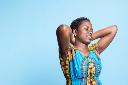 Casual African American woman relaxes in a studio, leaning back against blue background and spreading her arms. Young gorgeous and easygoing person unwinds in front of the camera.の写真素材