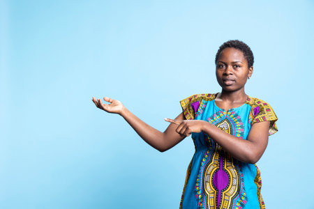 Confident woman in traditional ethnic costume creates advertisement, pointing at something on left or right sides. African american girl does web commercial ad in studio, natural beauty.の写真素材