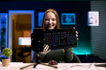 Joyous girl reviews keyboard, doing review for online streaming channel using mobile phone. Smiling media star excited to review computer peripherals, filming with smartphoneの写真素材