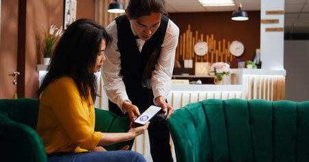 Tourist using nfc payment to buy coffee cup from hotel bar, employee bringing drink in lounge area. Asian woman relaxing in lobby, making mobile phone transaction at pos terminal. Handheld shot.の写真素材