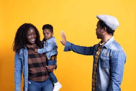 African american father waving hello at his little toddler and wife, meeting together against yellow background. Young black family posing in studio, dad having fun with small kid.の写真素材