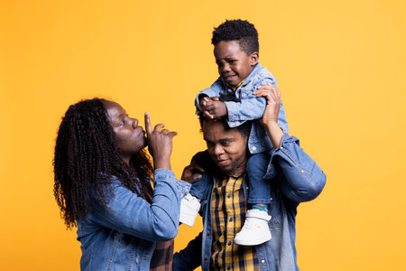 Parents calming down their infant son while he cries, his father is carrying him on his shoulders. Young mother showing hush sign to his toddler, trying to make him smile.の写真素材