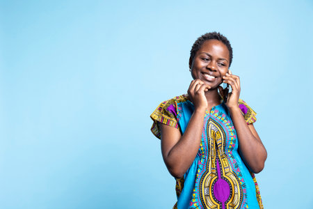 Excited woman using her mobile device to take calls, chuckling while she is engaging in a remote discussion with her friends against blue backdrop. In studio, african american person communicates.の写真素材