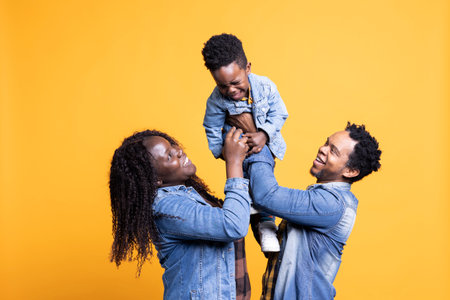 Loving african american parents lifting their cute small son in studio, posing in front of the camera. Proud happy couple bonding with their adorable toddler against yellow background.の写真素材