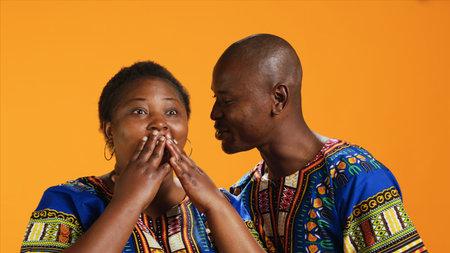Cheerful man sharing important secret with his wife, whispering in her ear words and telling gossip. Surprised woman leaning in and listening to confidential private information, bonding.の写真素材