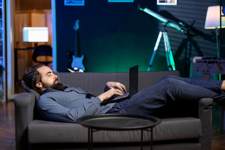 Freelancer lounged on couch working from home in apartment solving business related tasks on laptop. Remote employee laying down in neon lit living room, sending emails on notebookの写真素材