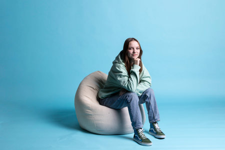 Portrait of happy girl sitting on beanbag armchair and smiling, isolated over blue studio background. Upbeat teenager seated on cozy bean bag, resting at home, having positive mood, studio backdropの写真素材