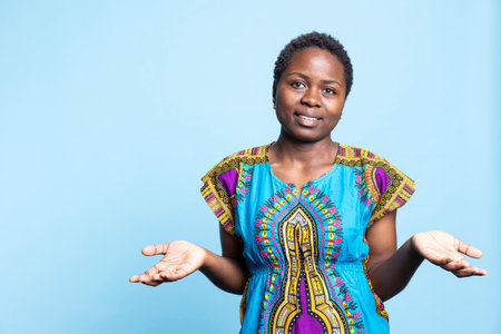 African american female model feeling doubtful and confused in studio, shrugging. Young adult feeling uncertain, traditional cultural clothing items.の写真素材