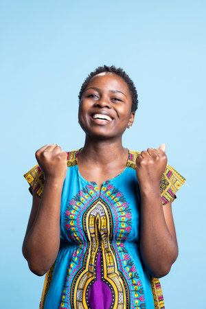 Excited African American girl giving an encouraging gesture while she looks at something in studio, ethnic costume. Natural beauty and energetic person posing against blue background.の写真素材