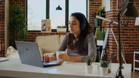 Indian woman drinking coffee in the morning and watching TV show at home using headphones. Matinal person waking up with hot beverage while enjoying television series on laptop, camera Aの写真素材