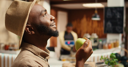 African american man smelling locally grown apples at supermarket, supporting nonpolluting small...