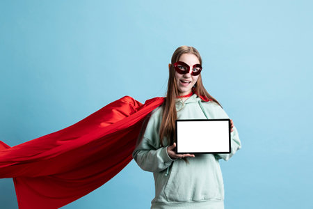 Joyful girl wearing superhero costume and mask for Halloween, using isolated screen tablet, studio background. Portrait of radiant person dressed as comic book hero holding mockup digital deviceの写真素材