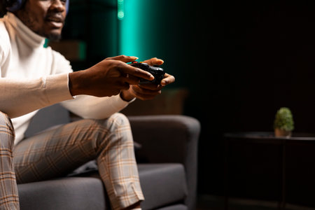 Gamer immersed in virtual world, holding controller in modern apartment, playing videogames. African american man enjoys gaming session from the comfort of his living roomの写真素材