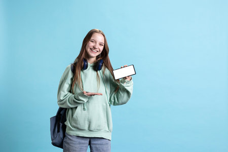 Jolly young girl smiling while presenting mobile phone with isolated screen display, studio background. Joyful woman has fun during promotion for mockup smartphone, studio backdropの写真素材