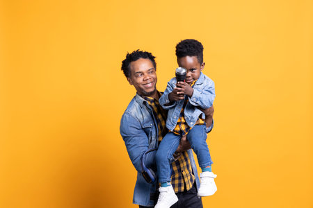 Joyful african american dad is carrying his toddler against yellow backdrop, curious small child playing with a flashlight in front of the camera. Enthusiastic father bonding with his son.の写真素材