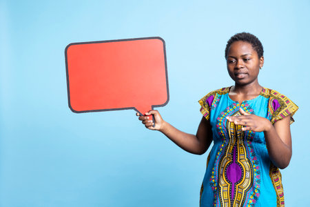 Young woman in traditional ethnic attire holds speech bubble sign, creating an ad commercial over blue background. Confident girl uses isolated mockup cardboard icon, natural beauty.の写真素材