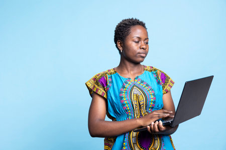 African american woman working on a laptop in front of the camera, using wireless pc to check social media and web pages. Young adult typing information online, internet connection in studio.の写真素材