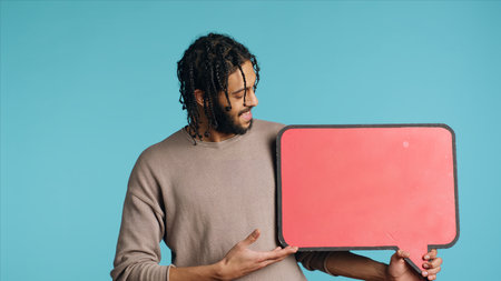 Joyful BIPOC man holding red speech bubble sign of empty space for text. Upbeat Middle Eastern...