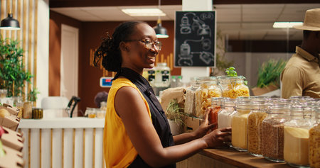 African american vendor arranging bulk products at local store, promoting zero waste concept for sustainable lifestyle. Merchant smiling and preparing to welcome clients at bio shop. Camera 2.の写真素材