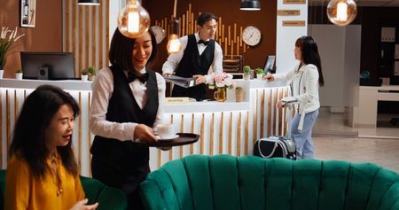 Asian woman asking receptionist about room reservation, arriving at hotel front desk. Staff team...