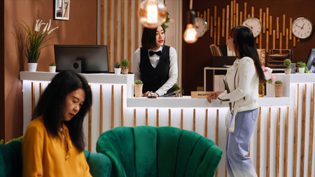 Asian client arriving at reception desk with trolley bags, staff asking tourist about her room reservation for check in process. Hotel concierge greeting guest at holiday resort, tourism concept.の写真素材