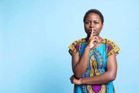 African american woman showing silence gesture on camera, presenting hush symbol to keep secret and be private. Female model doing mute sign over blue background, confidential information.の写真素材
