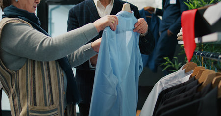 Wife searching for the right formal shirt for her husband, looking to expand wardrobe from sales at fashion boutique. Senior couple shopping for suits and elegant items, commercial activity. Camera B.の写真素材