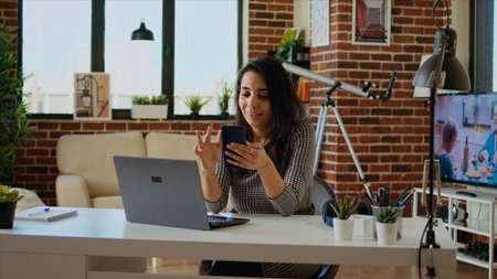 Teleworker checking her phone notifications at home in front of laptop, taking break from work. Woman in cozy apartment office taking pause from job tasks to read messages on smartphone, camera Aの写真素材