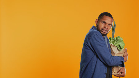 Vegan person protecting his eco friendly groceries bag, safeguarding his freshly harvested goods in studio. Serious man taking care of his bio organic belongings. Camera B.の写真素材
