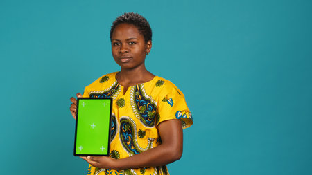 Gorgeous model presenting a tablet with green screen mockup, holding the gadget against blue background. Young charming woman in tribal clothing showing isolated display in studio. Camera A.の写真素材