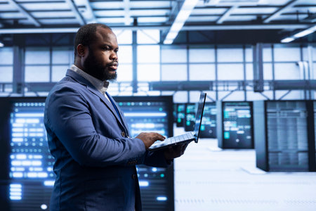 System administrator in data center implementing backup solutions to protect against data loss using laptop, ensuring efficiency. High tech facility worker resolving technical issues using notebookの写真素材