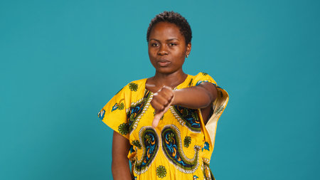 Young woman giving thumbs down in studio, showing her disappointment against blue background. Female model presenting a dislike symbol, express negativity and disapproval. Camera A.の写真素材
