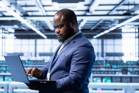 Computer scientist using laptop in data center with advanced tech, ensuring cybersecurity and efficient network management. African american expert in server hub optimizing data center with deviceの写真素材