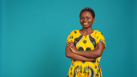 Portrait of young woman feeling confident in traditional native pattern attire, smiling and posing with arms crossed against blue background. Cheerful natural girl in dress. Camera B.の写真素材