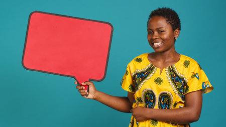 Woman in ethnic clothes holds a speech bubble carton board to create an ad commercial, presenting an isolated billboard icon in studio. Young adult posing for advertisement. Camera A.の写真素材