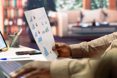 Business owner reviewing data infographics on papers at his home office setup, managing enterprise...