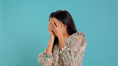 Fearful indian woman covering face with palms, worried about troubling peril, isolated over studio...