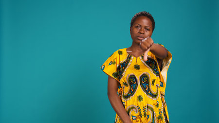 Dissatisfied person showing thumbs down symbol in studio, expressing her disapproval against blue background. Woman in ethnic tribal attire doing a dislike sign, disagreement. Camera B.の写真素材