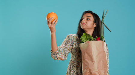 Indian woman with paper bag with vegetables and fruits testing quality, looking at orange, studio background. Vegetarian verifying groceries are ripe after buying them from zero waste shop, camera Aの写真素材