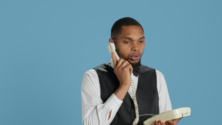 Bellboy concierge staff answering landline phone call to make reservations, discussing with clients about hotel rooms and amenities. Using telephone with cord to assist.の写真素材