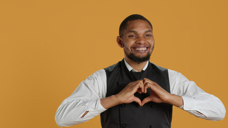Waiter in apron and uniform showing a heart shape sign in studio, expressing his sincere feelings and true love. Dining employee presents a romantic gesture, sweet confident man. Camera A.の写真素材