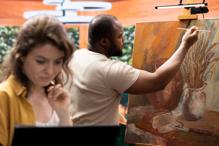 Woman searching for online inspiration while man is painting a masterpiece on canvas, doing research to find a model for their artwork. Team of artist collaborating on a painting.の写真素材