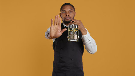 Waiter in uniform showing a stop sign with his palm raised in studio, expressing negative symbol and refusal about something. Showing rejection and being dissatisfied. Camera B.の写真素材