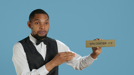 Hotel employee in tuxedo pointing people to the spa center using a wall sign, holding the indicator guide guests to resort facilities. Offering great services at the five star lodging. Camera A.の写真素材