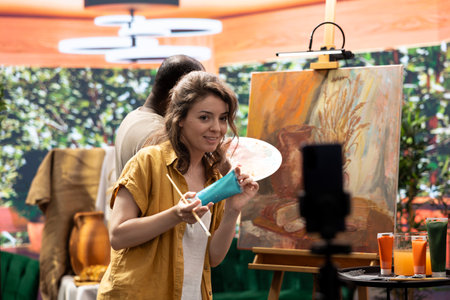 Female artist explaining the painting techniques used on online tutorial, recording a vlog for art class to teach people how to paint with watercolor on canvas. Team of painters do content creation.の写真素材