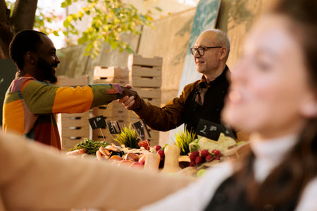 Selective focus of elderly vendor greeting african american client at greenmarket booth, offering fresh organic produce. Smiling multicultural individuals handshaking at farm food market stand.の写真素材