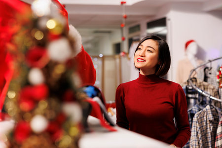 Santa claus man shows clothes to client in fashion boutique, person working in retail store and spreading holiday spirit with red costume. Asian woman asking man for help in buying items.の写真素材