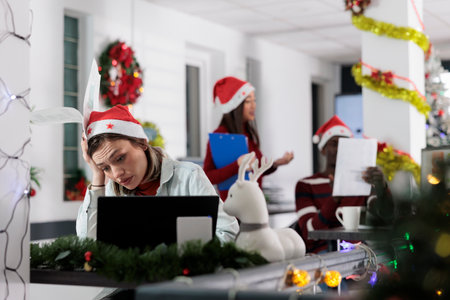 Office employee wearing Christmas hat feeling burnout while struggling to keep up with incoming company project deadline. Worker frustrated by paperwork overload during festive holiday seasonの写真素材