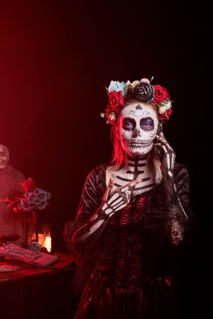 Woman talking on smartphone, celebrating Day of the Dead. Mysterious lady in telephone call, surrounded by funeral items during horror eventの写真素材
