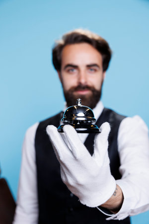 Porter with gloves holds service bell to show professional hotel concierge occupation, giving assistance to people. Young man employee in suit posing in studio, luxury hospitality industry.の写真素材